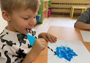 Zdjęcie przedstawia dzieci w trakcie rozdmuchiwania farby słomką